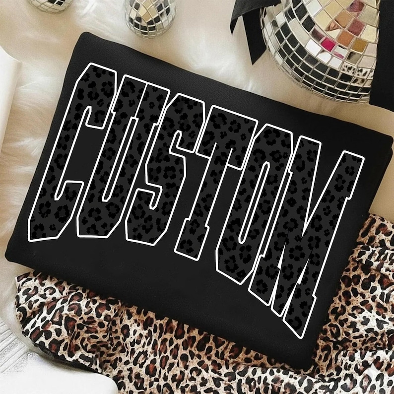 May include: Black t-shirt with the word "CUSTOM" in large, white outlined letters. The letters are filled with a leopard print pattern. The shirt is folded and resting on a leopard print fabric.