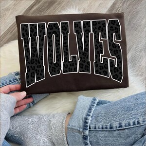 May include: A brown sweatshirt with the word "WOLVES" in large, block letters. The letters are black with a leopard print design and outlined in white. The sweatshirt is being held by a person wearing blue jeans.