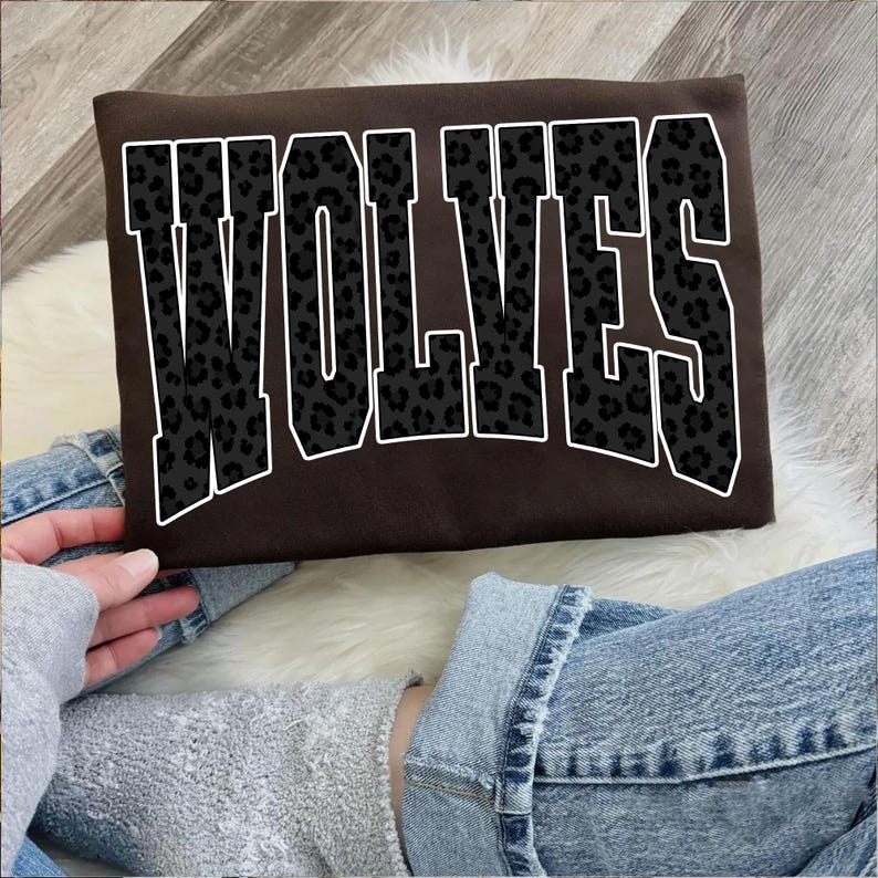 May include: A brown sweatshirt with the word "WOLVES" in large, block letters. The letters are black with a leopard print design and outlined in white. The sweatshirt is being held by a person wearing blue jeans.