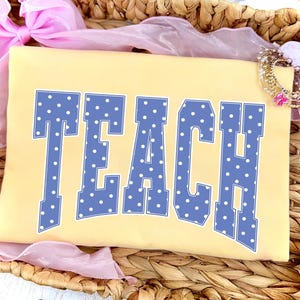 May include: A light yellow t-shirt with the word "TEACH" in large, blue, polka-dotted letters. The shirt is resting in a woven basket, with pink ribbon and a bracelet in the background. The shirt is a teacher-themed item.