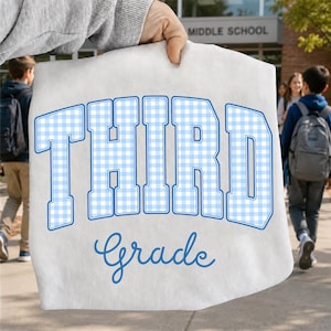 Könnte beinhalten: Ein weißes T-Shirt mit dem Wort THIRD in großen Blockbuchstaben mit einem hellblau-weißen Vichy-Karomuster, umrandet in Blau. Darunter ist das Wort Grade in einer blauen Schreibschrift in der Mitte des T-Shirts aufgedruckt.