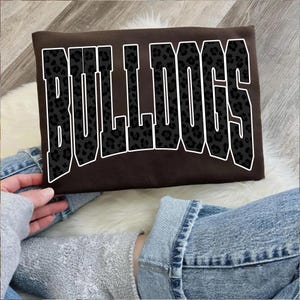 May include: A brown sweatshirt with the word "BULLDOGS" in large, bold, black leopard print letters outlined in white. The sweatshirt is rectangular and appears to be made of a soft material.