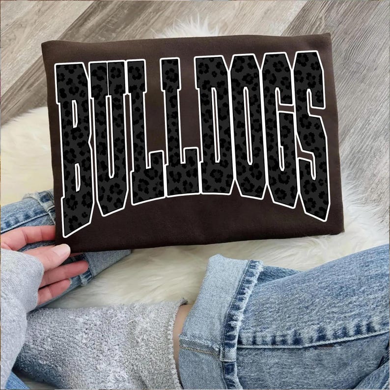 May include: A brown sweatshirt with the word "BULLDOGS" in large, bold, black leopard print letters outlined in white. The sweatshirt is rectangular and appears to be made of a soft material.