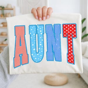 May include: A white t-shirt with the word "AUNT" in red and blue letters. The letters are decorated with gingham and star patterns, and outlined with a blue scalloped border. The shirt is held up by a person.