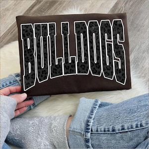 May include: A brown t-shirt with the word "BULLDOGS" in large, bold, black leopard print letters outlined in white. The shirt is displayed on a white fur-like surface, with denim jeans and a gray sweatshirt visible.