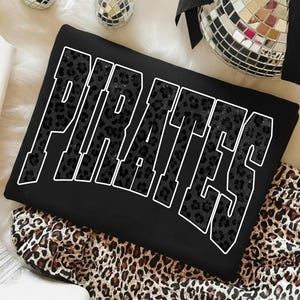 Könnte beinhalten: Schwarzes Sweatshirt mit dem Wort "PIRATES" in großen, weiß umrandeten Buchstaben. Die Buchstaben sind mit einem schwarzen Leopardenmuster gefüllt. Das Sweatshirt liegt auf einem Leopardenmuster-Stoff.