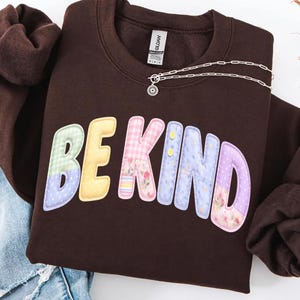 Könnte beinhalten: Ein braunes Sweatshirt mit den Worten "BE KIND" in bunten Buchstaben im Patchwork-Stil. Die Buchstaben sind in Gelb, Rosa, Blau und Lila gehalten. Eine silberne Halskette mit einem Blumenanhänger liegt auf dem Sweatshirt.