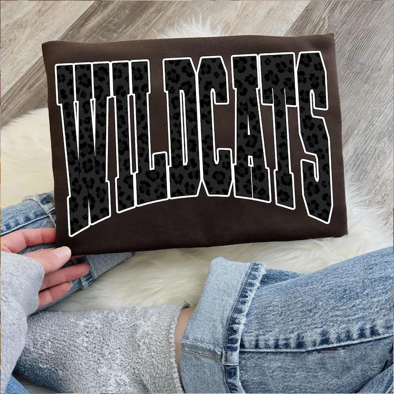 May include: A brown sweatshirt with the word "WILDCATS" in large, white-outlined letters. The letters are filled with a black leopard print pattern. The sweatshirt is being held by a person wearing a gray sweatshirt and blue jeans.