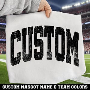 May include: A white t-shirt featuring the word CUSTOM printed in a large, black, distressed varsity-style font across the chest. The lettering has a weathered, cracked texture, and the shirt is held up against a blurred stadium background.