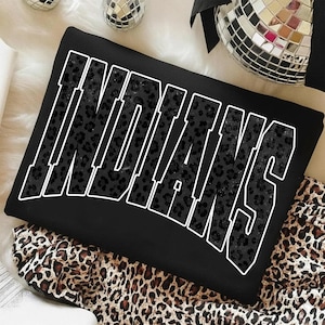 Könnte beinhalten: Schwarzes Sweatshirt mit dem Wort "INDIANS" in großen, weiß umrandeten Buchstaben. Die Buchstaben sind mit einem schwarzen Leopardenmuster gefüllt. Das Sweatshirt ist auf einem Leopardenmuster-Stoff gefaltet.
