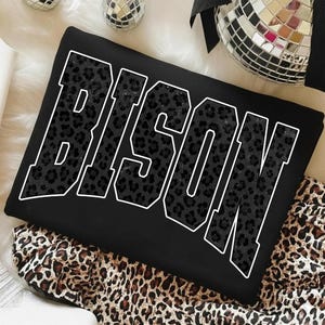 May include: Black sweatshirt with the word "BISON" in large, white-outlined letters. The letters are filled with a dark leopard print pattern. The sweatshirt is folded on a leopard print fabric.