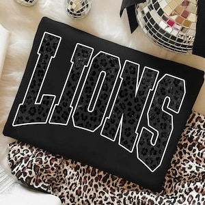 May include: Black sweatshirt with the word "LIONS" in large, white-outlined letters. The letters are filled with a dark leopard print pattern. The sweatshirt is folded on top of a leopard print fabric.