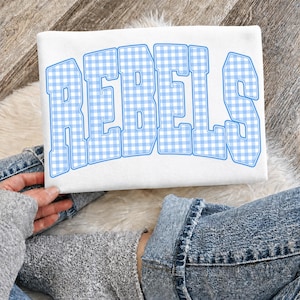 May include: White sweatshirt with the word "REBELS" in large, light blue and white gingham letters. The sweatshirt is being held by a person wearing blue jeans.