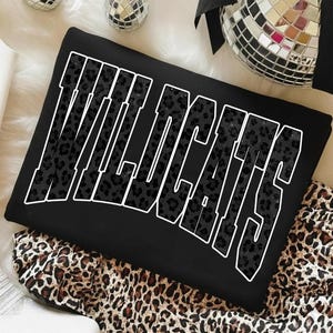 May include: Black sweatshirt with the word "WILDCATS" in large, white outlined letters. The letters are filled with a dark leopard print pattern. The sweatshirt is folded on top of a leopard print fabric.