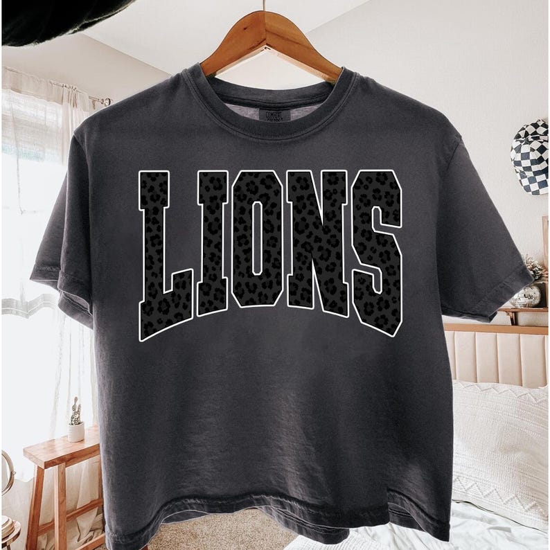 May include: A dark gray t-shirt with the word "LIONS" in large, white-outlined letters. The letters are filled with a leopard print pattern. The shirt is hanging on a wooden hanger.