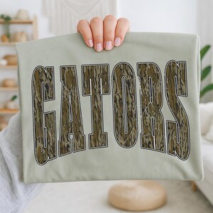 Pode incluir: Uma t-shirt verde sálvia claro com a palavra GATORS em letras maiúsculas grandes e arqueadas. O texto tem uma aparência desgastada e envelhecida, preenchido com um padrão de camuflagem castanho, verde e preto com um contorno branco fino.