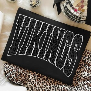 Puede incluir: Sudadera negra con la palabra "VIKINGS" en letras grandes con contorno blanco. Las letras están rellenas con un estampado de leopardo oscuro. La sudadera está sobre una tela con estampado de leopardo.