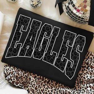 May include: Black sweatshirt with the word "EAGLES" in large, white-outlined letters. The letters are filled with a black leopard print pattern. The sweatshirt is folded on a leopard print fabric.