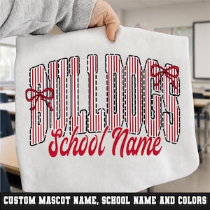May include: A white fabric item featuring the word BULLDOGS in large, red and white vertical striped block letters with black outlines and two red bows. Below, the text School Name is printed in a red, cursive script font on the white surface.