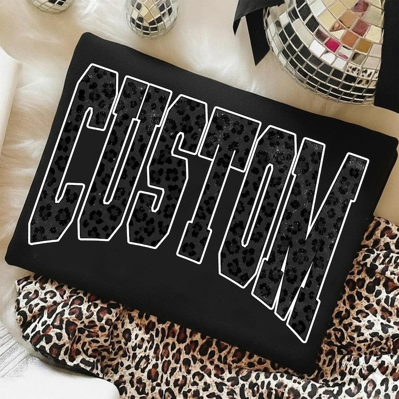 May include: Black sweatshirt with the word "CUSTOM" in large, white-outlined letters. The letters are filled with a black leopard print pattern. The sweatshirt is folded on top of a leopard print fabric.