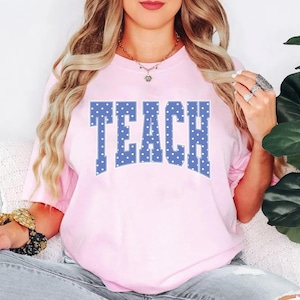 May include: A light pink t-shirt with the word "TEACH" in large, blue, polka-dotted letters. The shirt has short sleeves and a crew neck. The overall style is casual and comfortable.