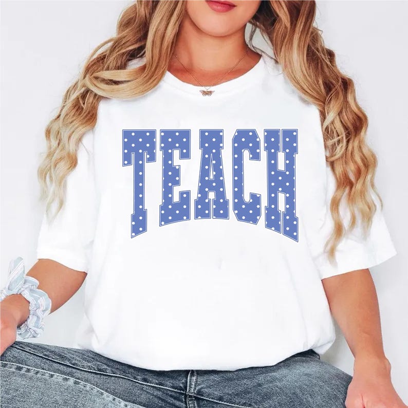 May include: White t-shirt with the word "TEACH" in large, blue, polka-dotted letters. The shirt is a casual style, suitable for everyday wear. The design is centered on the front of the shirt.