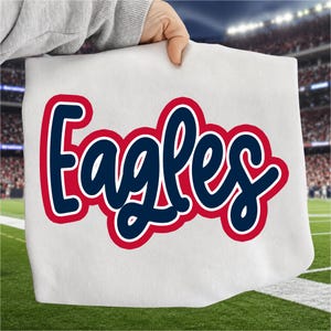 Könnte beinhalten: Ein weißes T-Shirt mit dem Wort Eagles in einer dunkelblauen Schreibschrift mit einer dicken roten Umrandung. Der Schriftzug ist mittig auf dem Stoff platziert und zeigt einen kräftigen, abgerundeten Stil, der sich vom weißen Hintergrund abhebt.