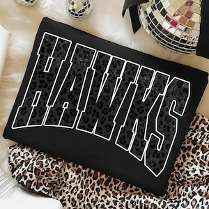 May include: Black sweatshirt with the word "HAWKS" in large, white outlined letters. The letters are filled with a black leopard print pattern. The sweatshirt is on a leopard print fabric.