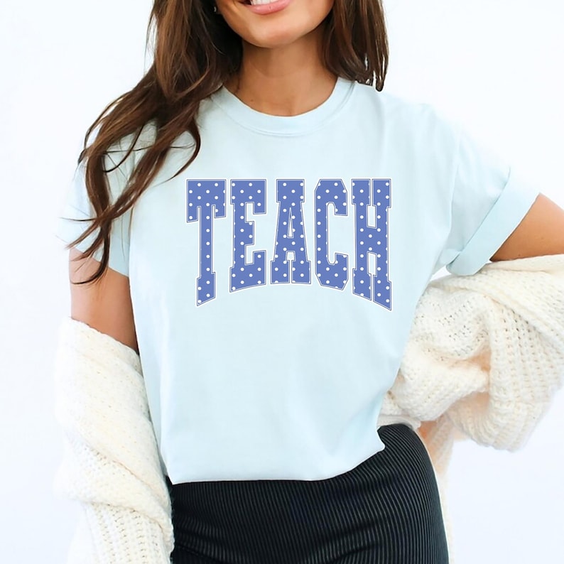 May include: Light blue t-shirt with the word "TEACH" in large, blue, polka-dotted letters. The shirt has short sleeves and a crew neck. The shirt is made of a soft material.