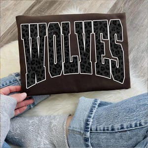 May include: A brown sweatshirt with the word "WOLVES" in large, block letters. The letters have a black leopard print pattern and are outlined in white. The sweatshirt is being held by a person wearing a gray sweatshirt and blue jeans.