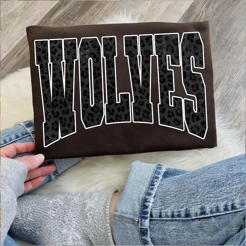 May include: A brown sweatshirt with the word "WOLVES" in large, block letters. The letters have a black leopard print pattern and are outlined in white. The sweatshirt is being held by a person wearing a gray sweatshirt and blue jeans.
