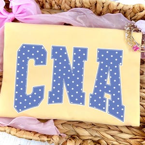 Könnte beinhalten: Ein hellgelbes T-Shirt mit den Buchstaben "CNA" in Blau mit weißen Punkten. Das Shirt wird in einem geflochtenen Korb mit rosa Schleife und einem dekorativen Armband präsentiert.