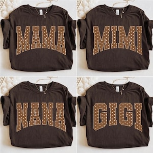 Könnte beinhalten: Eine Kollektion aus vier dunkelbraunen T-Shirts mit kurzen Ärmeln, die jeweils eine große, geschwungene Schrift im College-Stil mit weißen Punkten aufweisen. Die Shirts zeigen die Wörter MAMA, MIMI, NANA und GIGI in fetter Blockschrift.