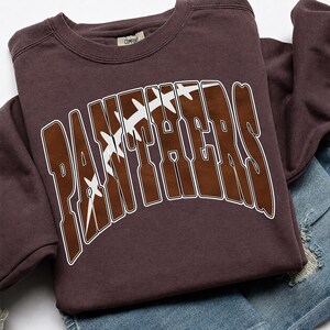 May include: A dark brown crewneck sweatshirt featuring the word PANTHERS in large, arched, brown block letters with a white outline. The letters are filled with a graphic of a brown football with white laces running diagonally across the text.