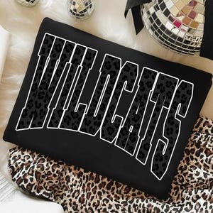May include: Black sweatshirt with the word "WILDCATS" in large, white outlined letters. The letters are filled with a leopard print pattern. The sweatshirt is folded on top of a leopard print fabric.