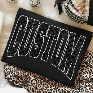 Könnte beinhalten: Schwarzes Sweatshirt mit dem Wort "CUSTOM" in großen, weiß umrandeten Buchstaben. Die Buchstaben sind mit einem Schachbrettmuster und einem Leopardenmuster gefüllt. Das Sweatshirt liegt auf einem Leopardenmuster-Stoff.