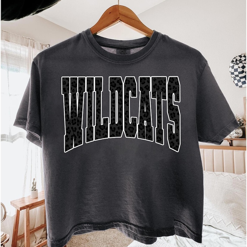 May include: A charcoal gray t-shirt with the word "WILDCATS" in large, bold letters across the front. The letters are filled with a black leopard print pattern and outlined in white. The shirt is hanging on a wooden hanger.