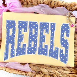 May include: A light yellow t-shirt with the word "REBELS" in large, blue, polka-dotted letters. The shirt is folded and resting in a woven basket, with pink ribbon and a bracelet in the background.