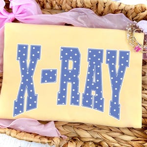 May include: A light yellow t-shirt with the word "X-RAY" in large, blue, polka-dotted letters. The shirt is resting in a woven basket, with pink fabric and a decorative bracelet in the background.