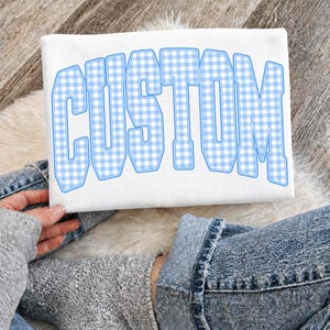 May include: White sweatshirt with the word "CUSTOM" in large, light blue gingham letters. The sweatshirt is displayed on a person's lap, with denim jeans and a gray sweater visible.