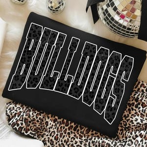 May include: Black sweatshirt with the word "BULLDOGS" in large, white-outlined letters. The letters are filled with a dark leopard print pattern. The sweatshirt is folded on a leopard print fabric.
