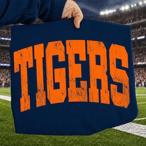 Pode incluir: Um tecido azul marinho com a palavra "TIGERS" em letras laranja desgastadas. A imagem é exibida em frente a um fundo desfocado de um estádio de futebol com gramado verde e linhas brancas.