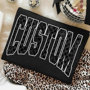 Könnte beinhalten: Schwarzes Sweatshirt mit dem Wort "CUSTOM" in großen, weiß umrandeten Buchstaben. Die Buchstaben sind mit einem Schachbrettmuster und einem Leopardenmuster gefüllt. Das Sweatshirt liegt auf einem Leopardenmuster-Stoff.