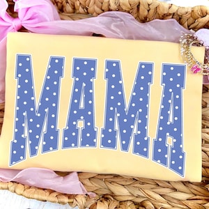 May include: A light yellow t-shirt with the word "MAMA" in large, blue, polka-dotted letters. The shirt is displayed in a woven basket with pink ribbon and jewelry accents.