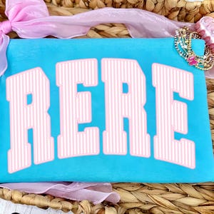 Puede incluir: Una bolsa turquesa con la palabra "RERE" en letras grandes, rosas y rayadas. Una pulsera dorada y turquesa con un dije de mariposa rosa está en la esquina superior derecha. Un lazo y una cinta rosas están en el fondo.