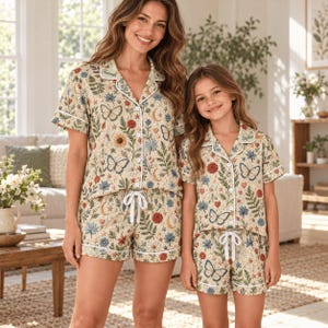 May include: Matching pajama sets featuring a cream-colored fabric with a botanical print of butterflies, flowers, ferns, and crescent moons. The short-sleeved button-down shirts have white piping and collars embroidered with the names Hannah and Sophie.