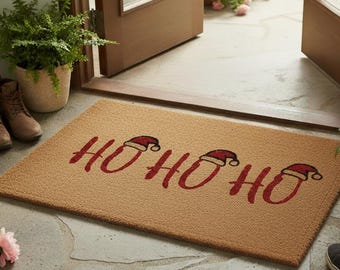 Op de afbeelding: Een rechthoekige deurmat met de woorden "HO HO HO" in rood schrift, elke "O" bekroond met een kerstmuts. De mat heeft een natuurlijke bruine kleur en is voor een deuropening geplaatst. Een paar bruine laarzen en sneakers staan in de buurt.