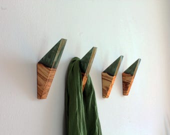 May include: Four wooden wall hooks with a geometric design. The hooks are made of olive wood and dark green resin. A green scarf hangs from one of the hooks. The hooks are mounted on a white wall.