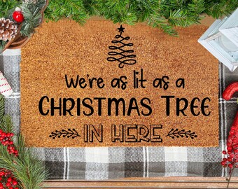 May include: A brown rectangular doormat with the text "We're as lit as a CHRISTMAS TREE IN HERE" in black. The mat features a Christmas tree design. The image is framed by Christmas decorations, including fir branches and berries.