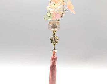 May include: A decorative keychain featuring a small bouquet of artificial flowers in shades of pink, white, and green. The flowers are arranged in a clear, glass-like vase, accented with gold-colored metal and a pink tassel. The keychain is suspended from a gold chain.
